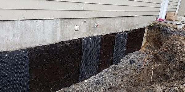 Réparation des fondations extérieures : une membrane de drainage noire est installée sur le mur de fondation en béton. La terre est excavée autour du mur.