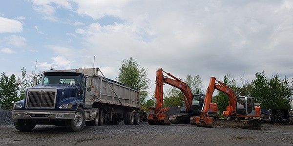 Un camion-benne et deux excavatrices orange sur un terrain gravillonné sous un ciel nuageux.