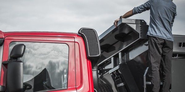 Une personne debout sur la benne d'un camion, tendant la main vers le compartiment métallique noir ouvert, à côté du camion rouge.