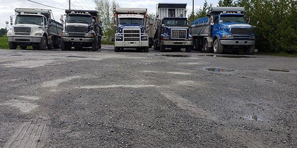 Cinq camions-bennes sont stationnés sur un terrain gravillonné. Les camions sont de couleurs différentes.