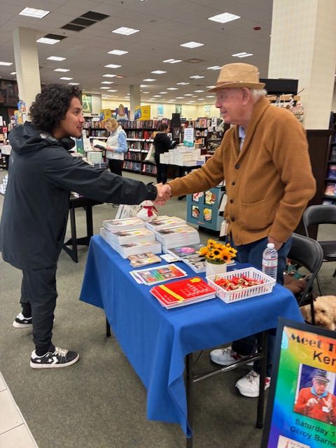 Two people shake hands at a table of books in a bookstore.