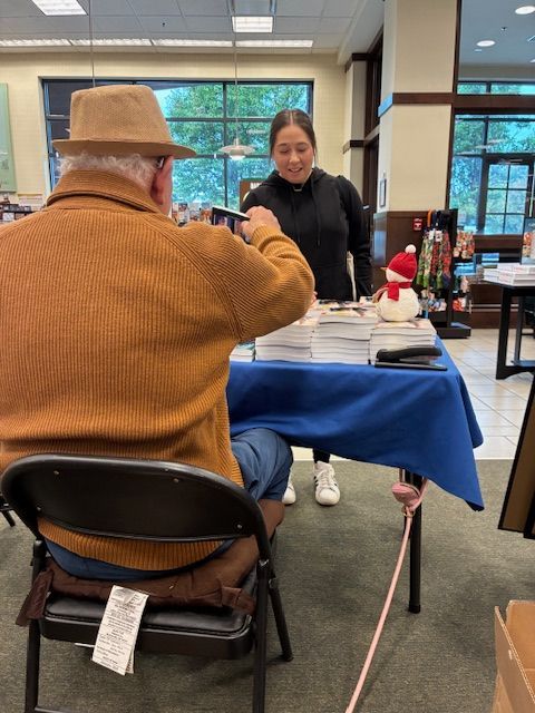 Man seated at a table, receiving assistance from a store employee at a book signing.