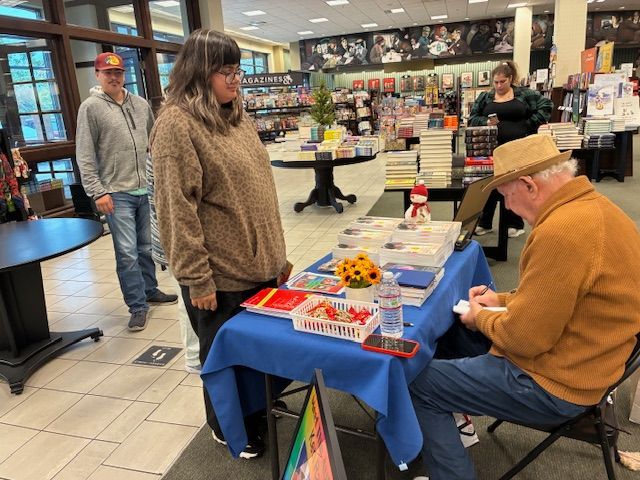 Person signing books at a table in a bookstore. A customer watches. Others browse books nearby.
