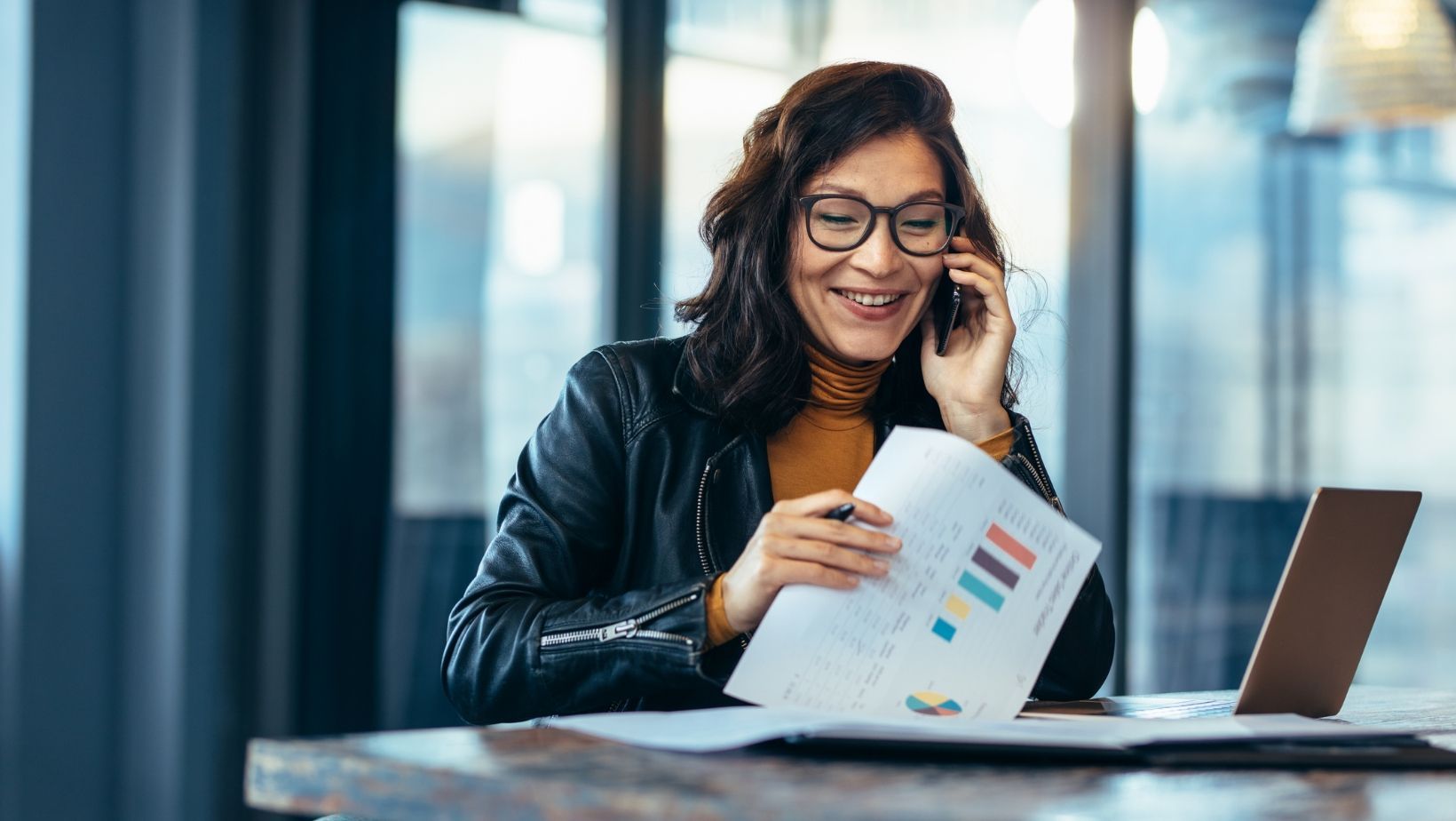A woman is sitting at a table talking on a cell phone while holding a piece of paper.
