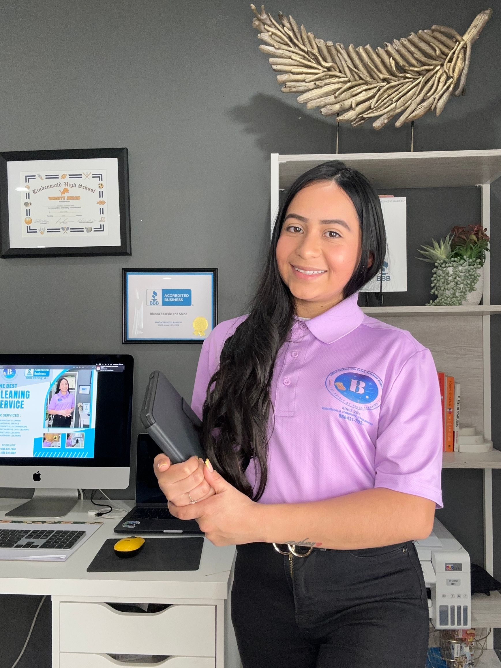 A smiling person in a light purple polo shirt stands in an office holding a tablet, with a computer and shelves nearby.