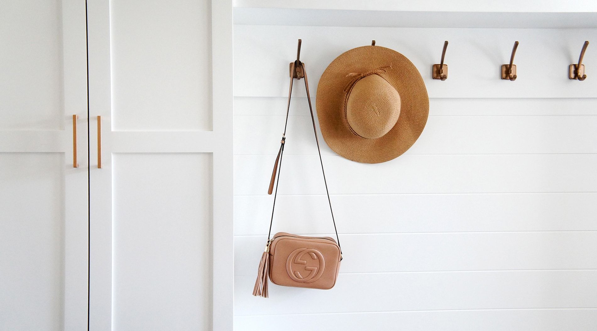 White wall with hooks holding a straw hat and a small purse. A white cabinet is on the left.
