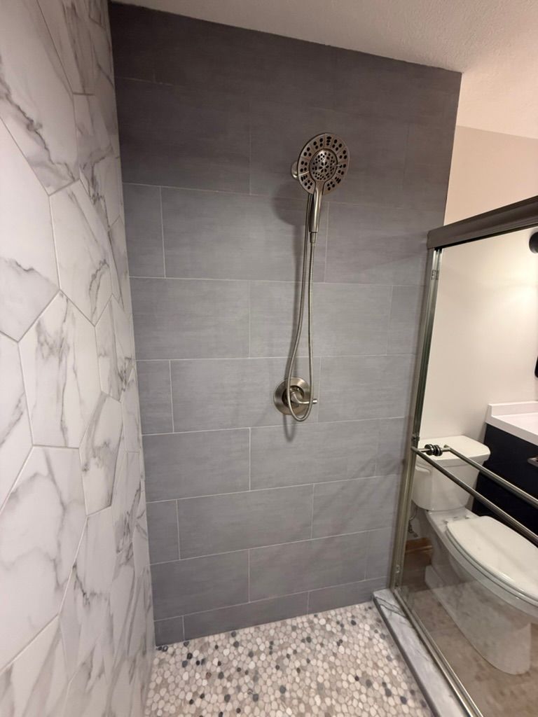 Small bathroom shower with gray tile walls, marble-look side wall, and pebble-pattern floor beside a vanity.