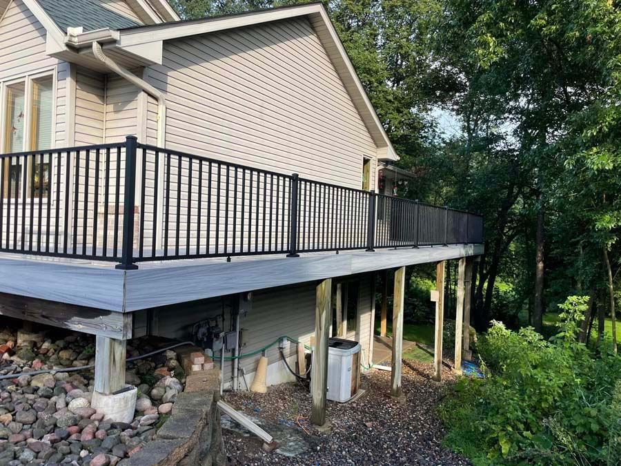 Raised wooden deck attached to a beige house, with black railings and stone support posts beside trees