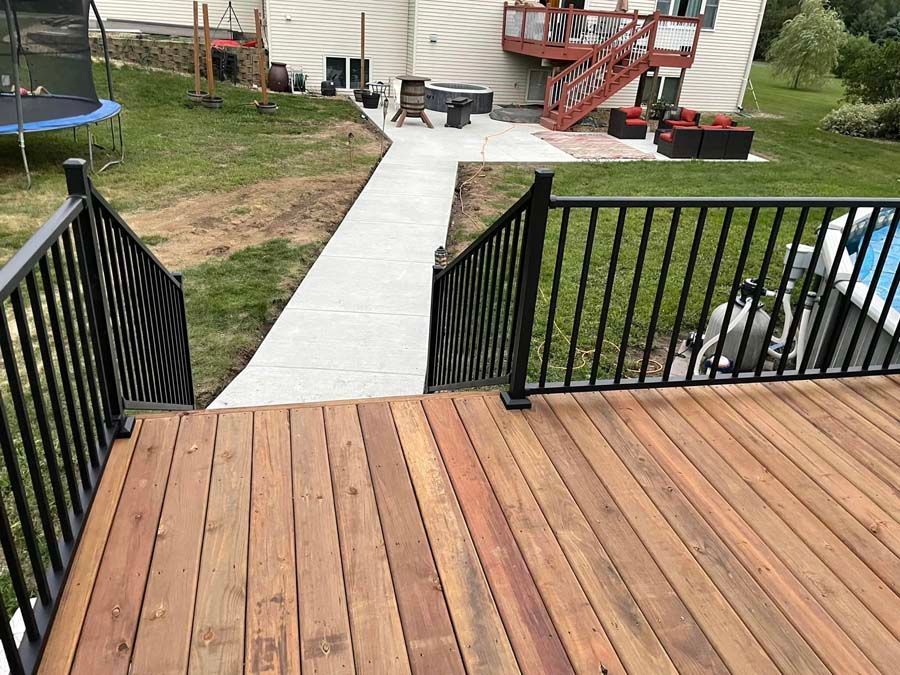Wooden deck steps leading to a concrete walkway beside black railings and a backyard patio area.