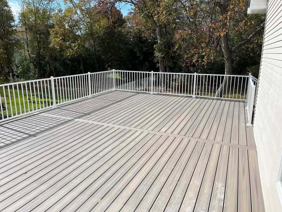Wooden deck with white railing beside a house, overlooking trees and a grassy yard.