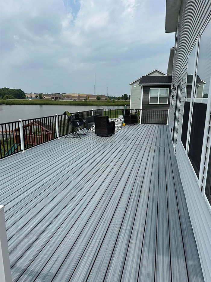 Rooftop deck with gray flooring, black railing, water view, and a small white building under cloudy skies