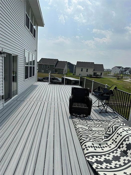 Backyard deck with black outdoor chair, patterned rug, and neighboring houses under a bright sky