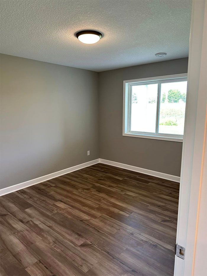 Empty beige room with wood-look flooring, ceiling light, and a window showing daylight.
