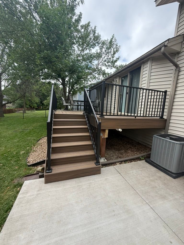 Concrete patio with wooden stairs leading to a raised deck beside a house and trees.