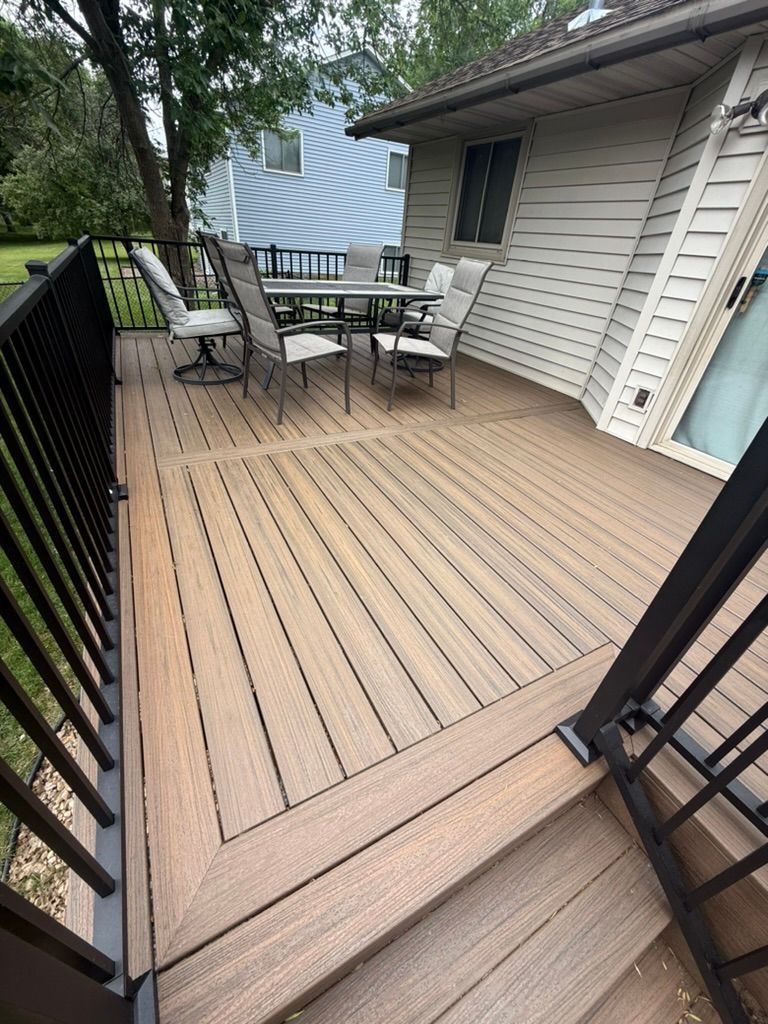 Wooden deck with patio table and chairs beside a house, viewed through a black gate and fence.