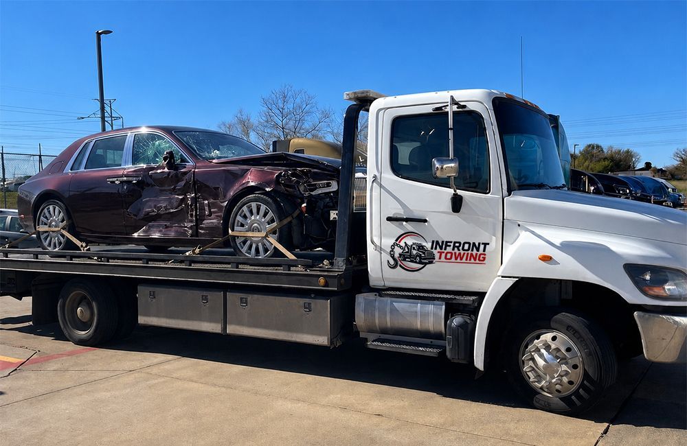 Tow truck carrying a damaged maroon sedan on a flatbed in a sunny lot