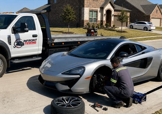 Mechanic changing a tire on a silver sports car in a suburban driveway beside a service truck