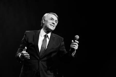 Een man in pak en stropdas zingt op een podium in een microfoon.
