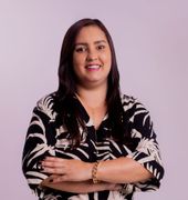 Mulher sorridente com os braços cruzados, vestindo uma blusa estampada em preto e branco, contra um fundo lilás claro.