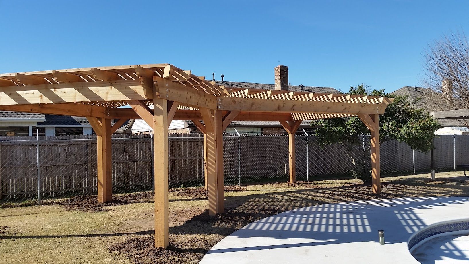 Wooden pergola over a backyard patio beside a fenced yard under a clear blue sky