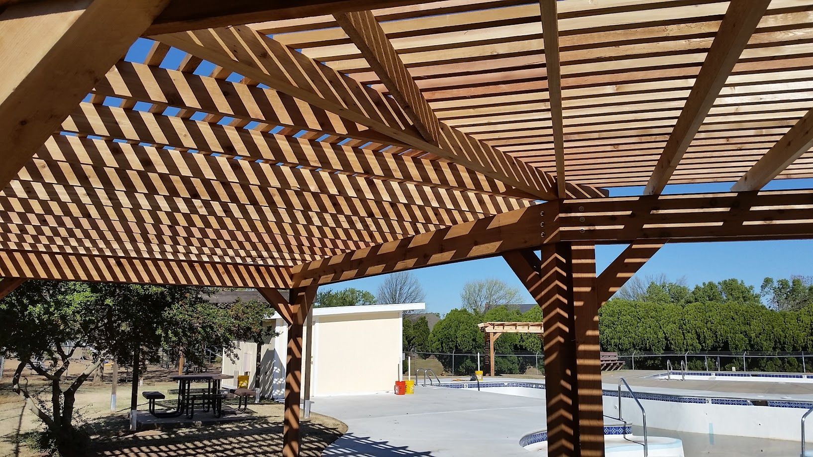 Wooden pergola shading an outdoor pool area with lounge chairs and trees in the background
