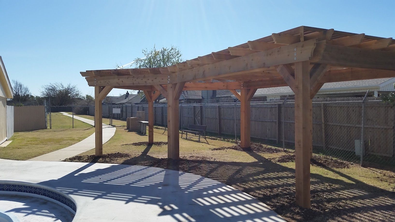 Wooden pergola beside a swimming pool in a fenced backyard under bright sunlight