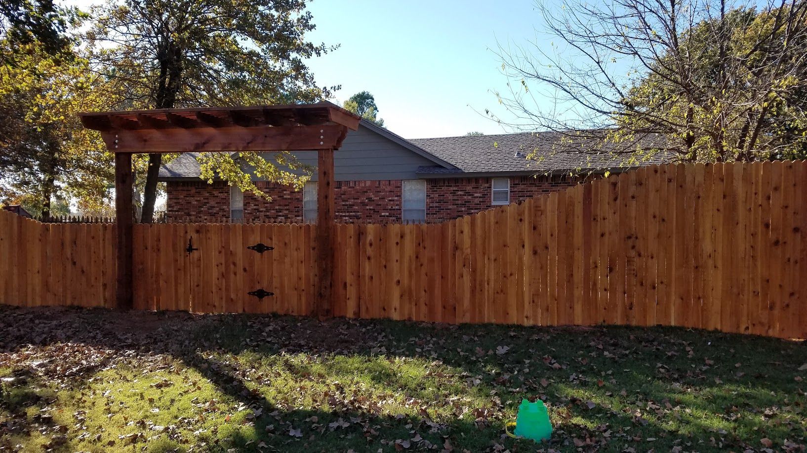 Backyard with a tall wooden fence, pergola, house roof, and trees in daylight.