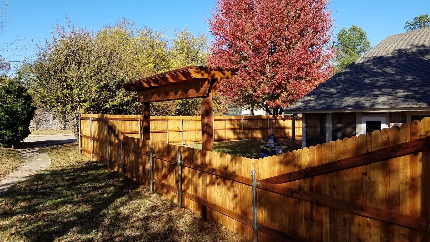 Wooden fence and gate in a sunny yard with a red autumn tree beside a house
