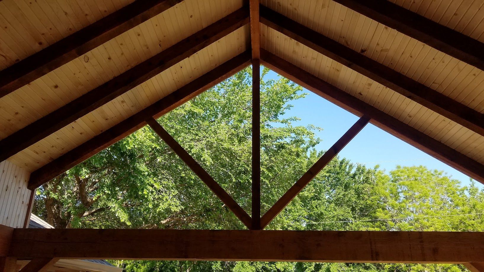 Wooden roof trusses framing a view of green trees and blue sky outdoors
