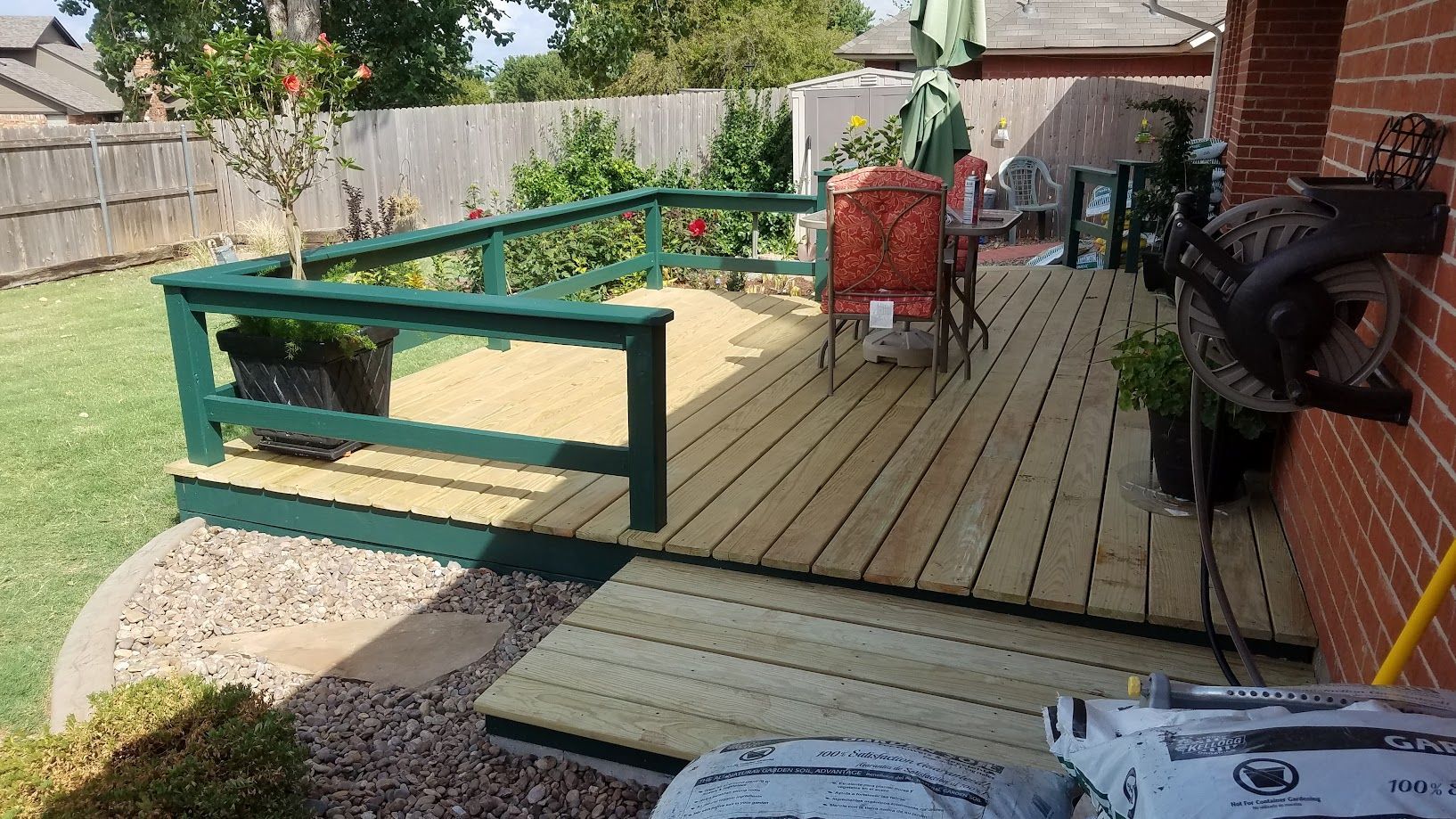 Backyard deck with green railing, patio chairs, grill, and a brick house wall in sunlight