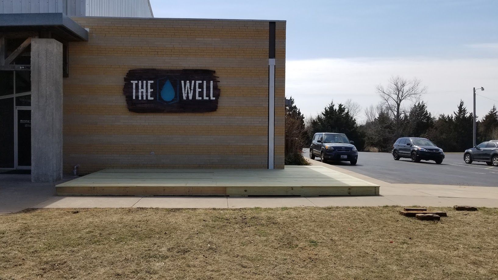 Building exterior with “The Well” sign, concrete sidewalk, and parked cars in a lot.