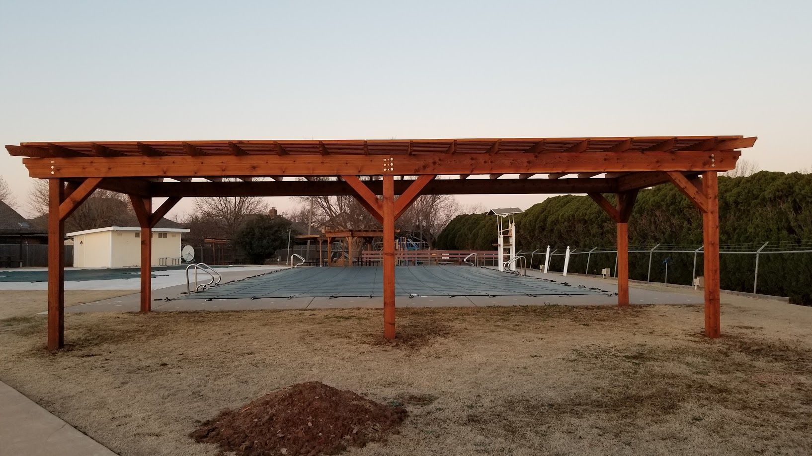 Wooden pergola with benches over a gravel area at dusk, with trees and a small structure in the background
