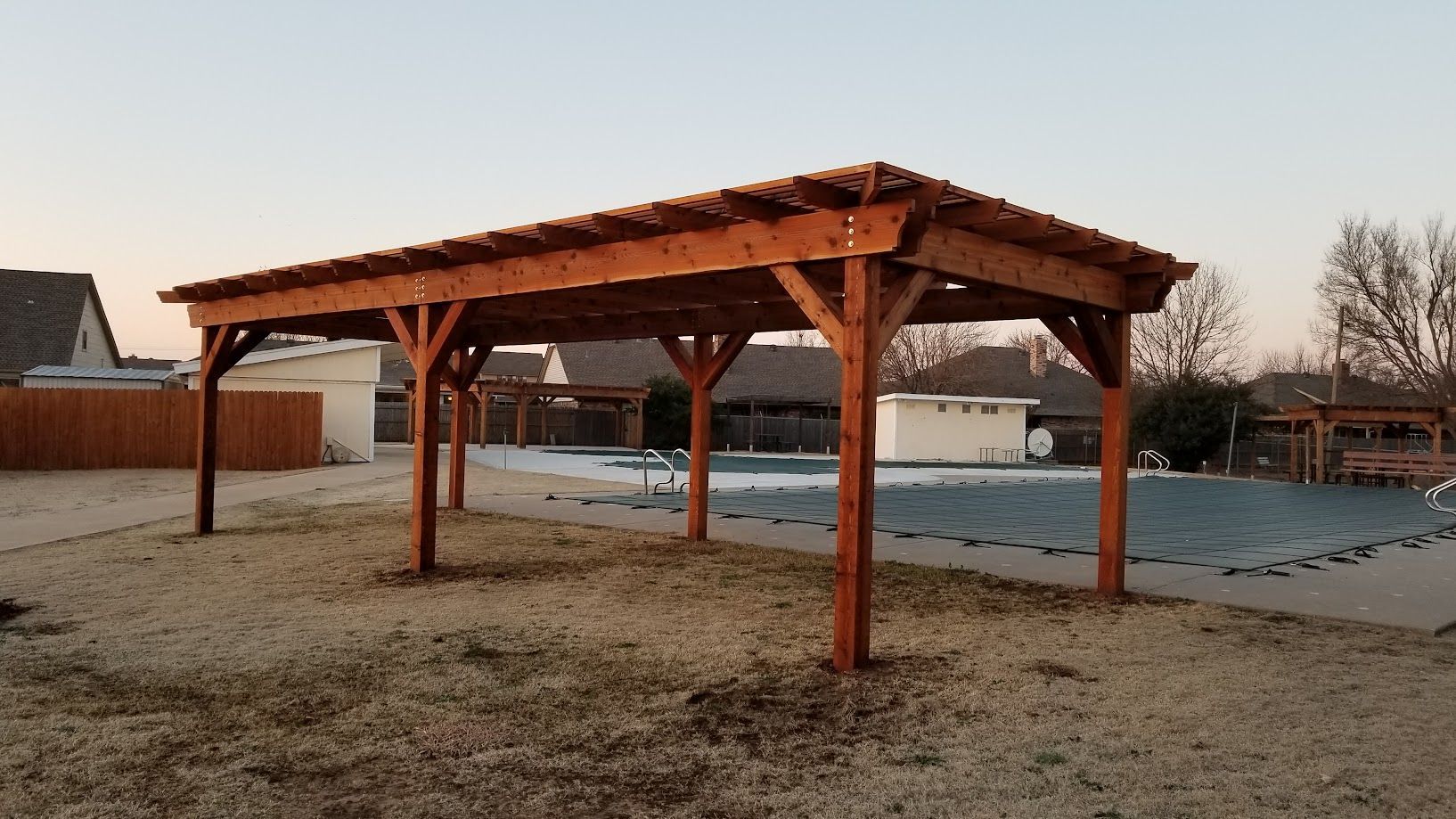 Wooden pergola in a snowy yard at sunset, with houses and a fence in the background