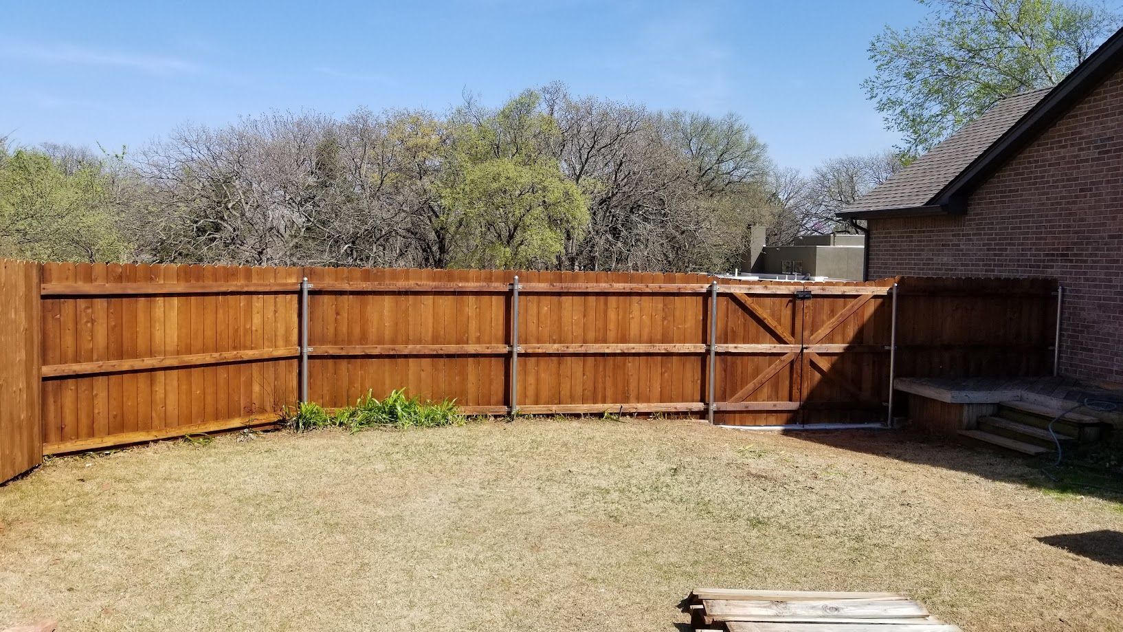 Wooden fenced backyard with dry grass and a brick house wall on the right.