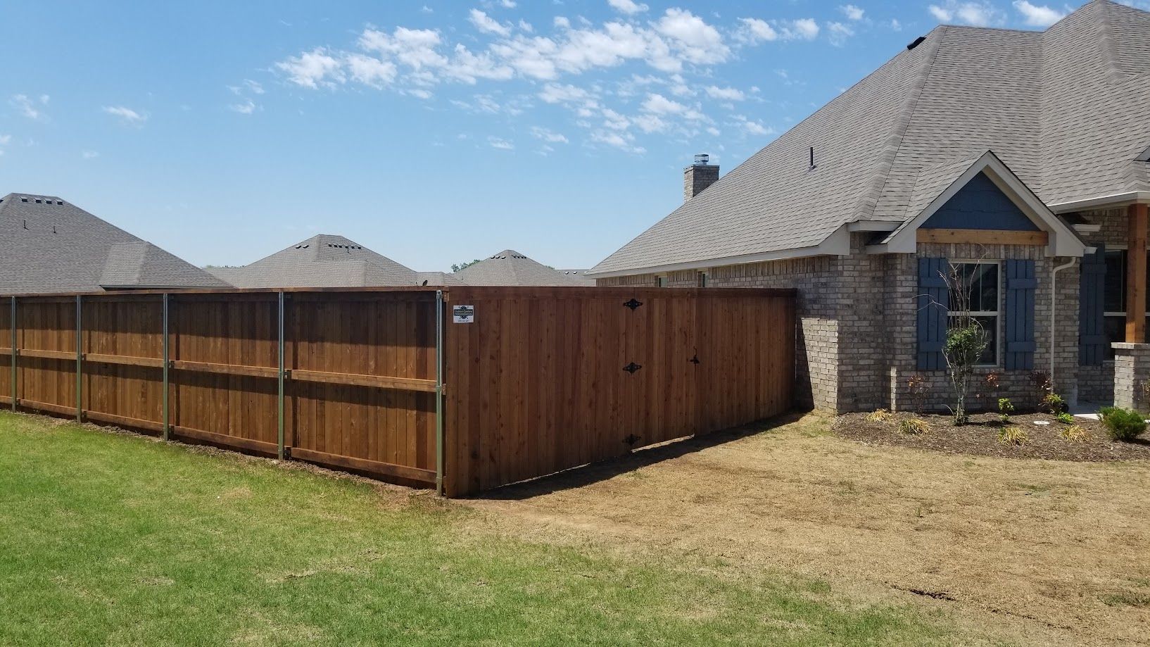 Suburban brick house with wooden privacy fence and dry lawn under a blue sky