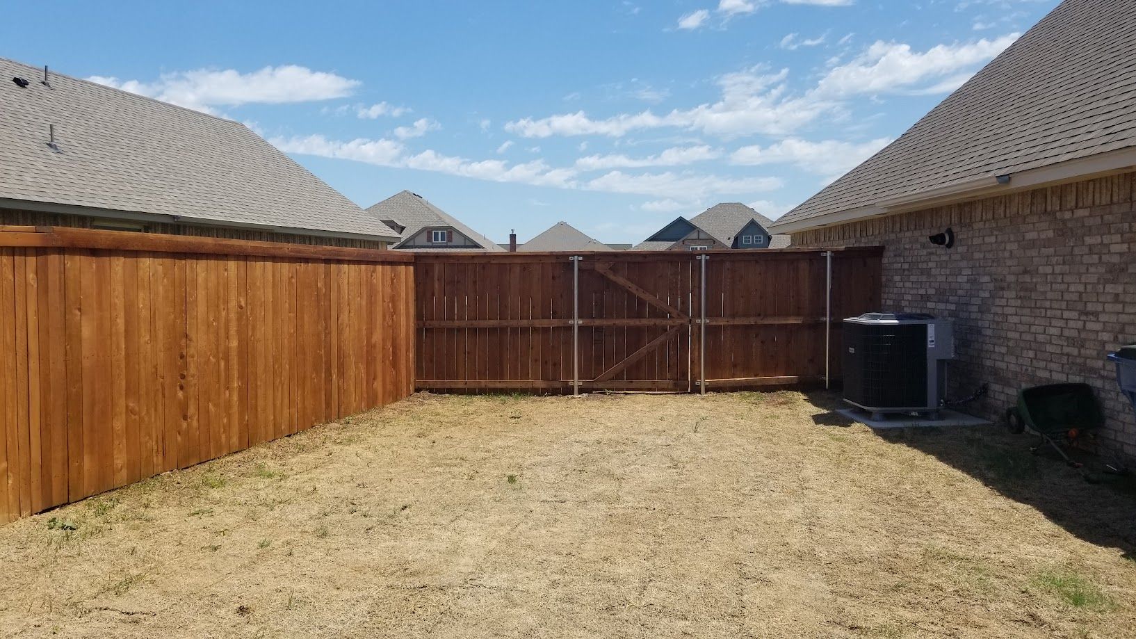 Backyard with dry grass, wooden fence, brick house walls, and a closed gate under a blue sky