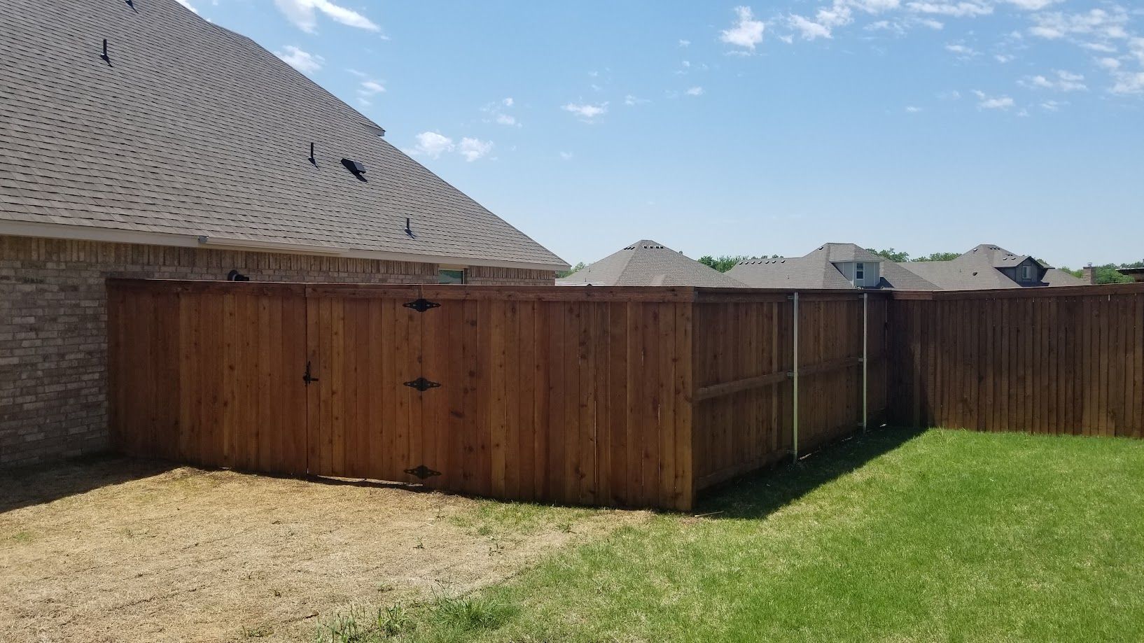 Wooden backyard fence and gate beside a brick house, with green grass and a blue sky.