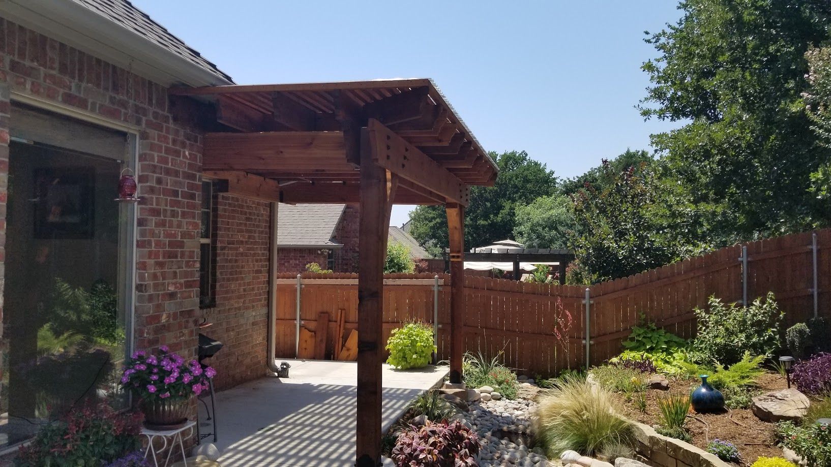 Backyard patio with brick house, wooden pergola, colorful flowers, and fence under bright sunlight