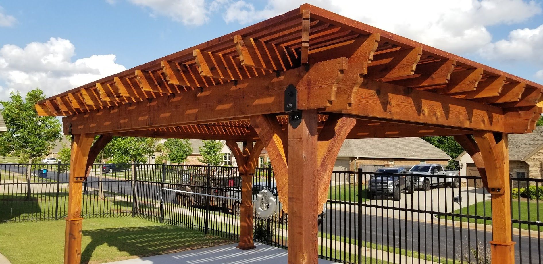 Wooden pergola structure beside a fenced walkway in a grassy park under a blue sky