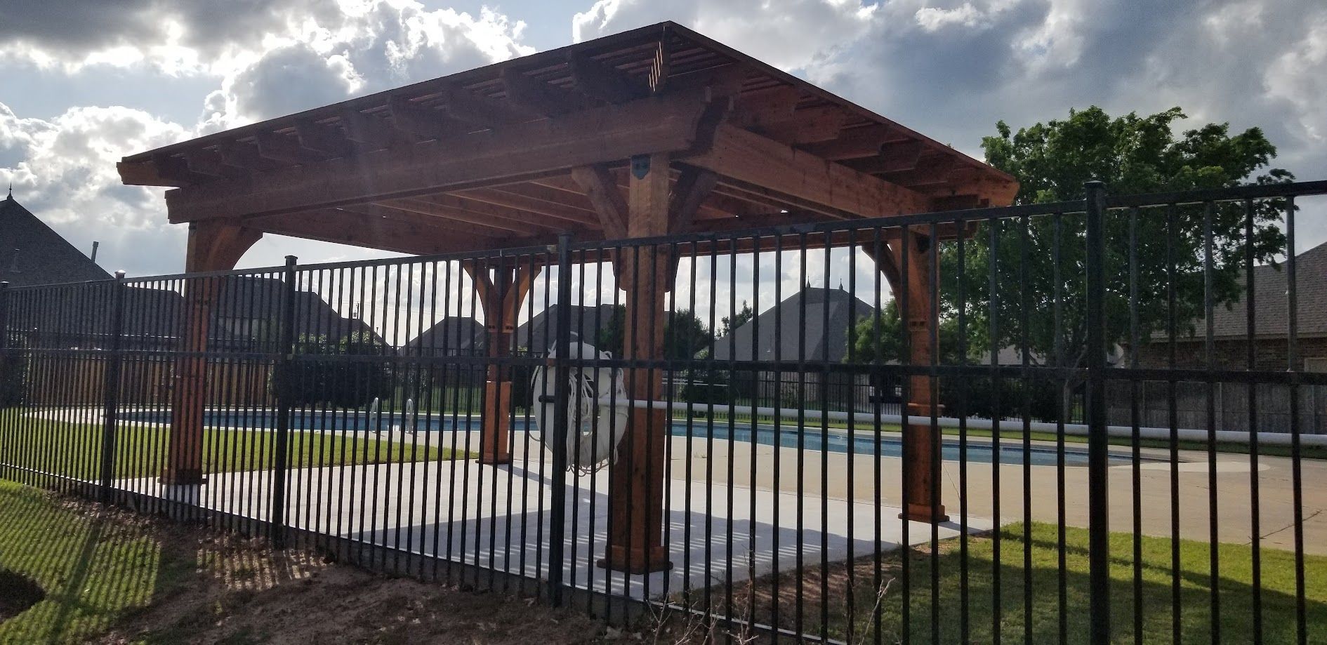 Covered outdoor pavilion with wooden roof and benches behind a black metal fence under a cloudy sky
