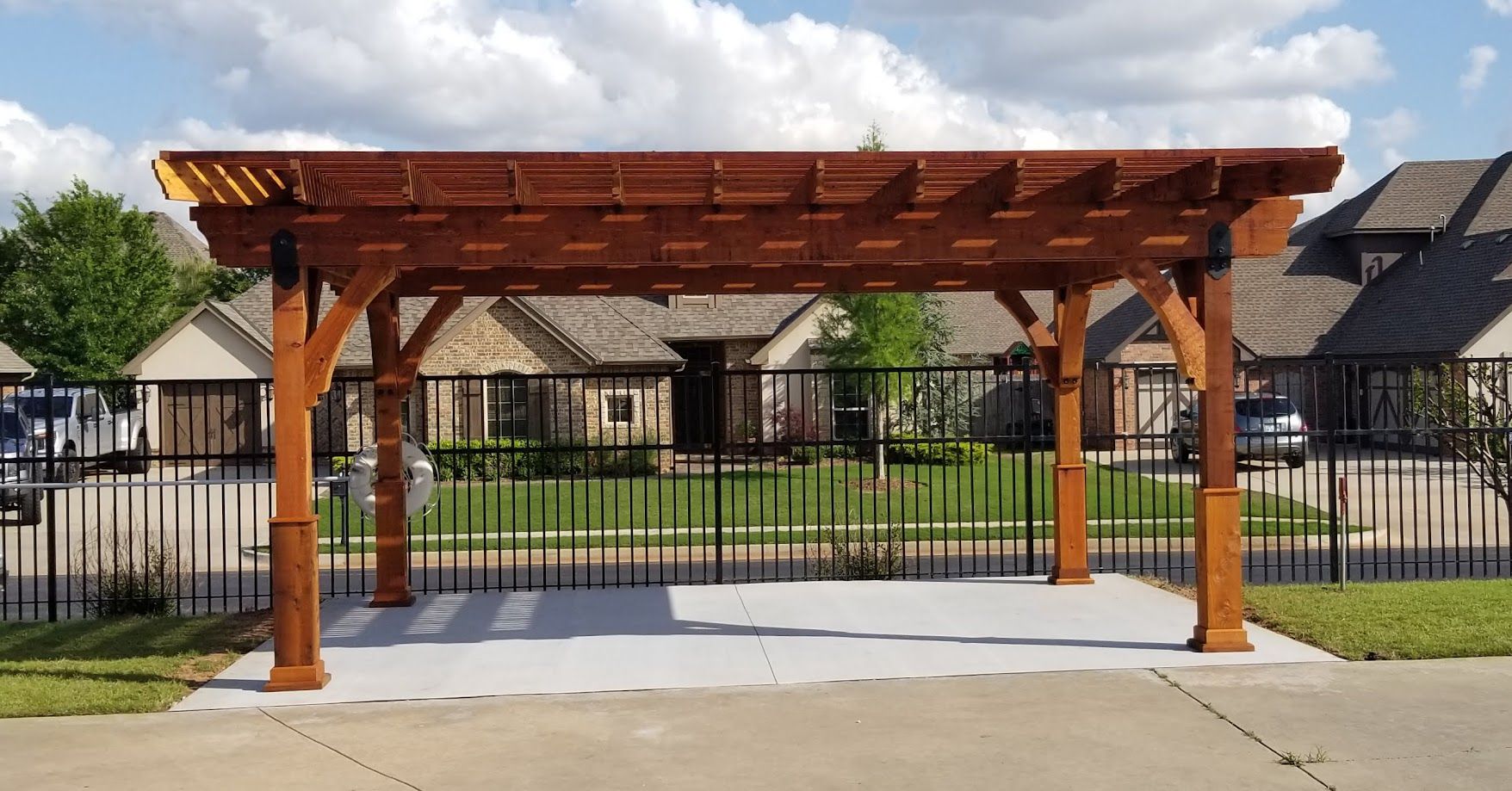 Wooden pergola shelter on a sidewalk in front of a fenced residential area, under a partly cloudy sky