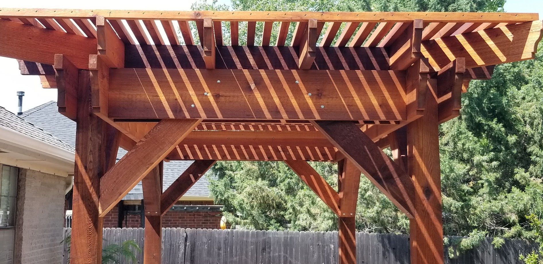 Wooden pergola with slatted roof and benches in a backyard, lit by sunlight