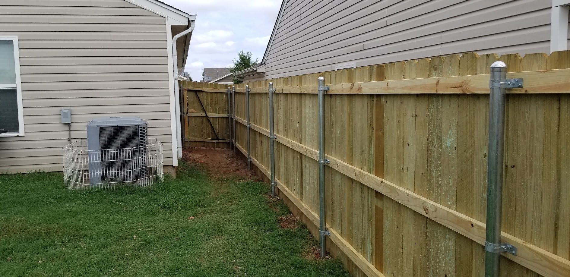 Narrow grassy side yard between a house and wooden fence, with an AC unit near the house.