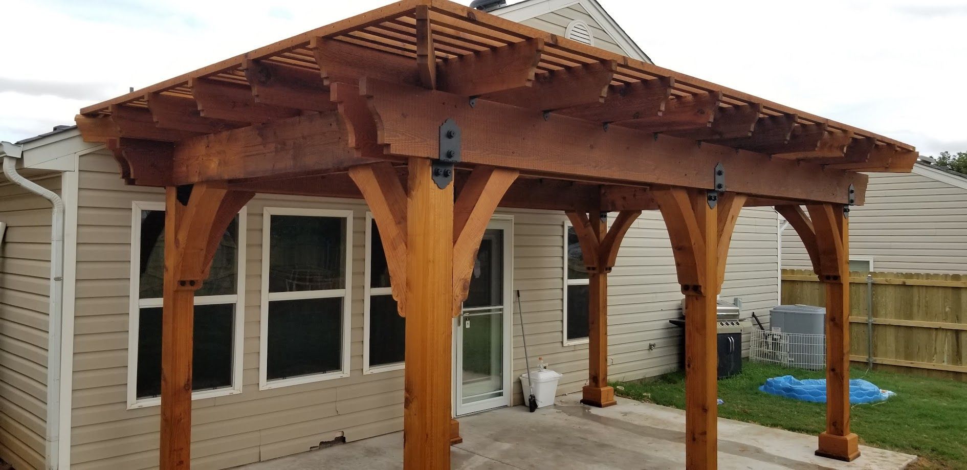 Wooden pergola attached to a beige house with white-trimmed windows and a covered patio.