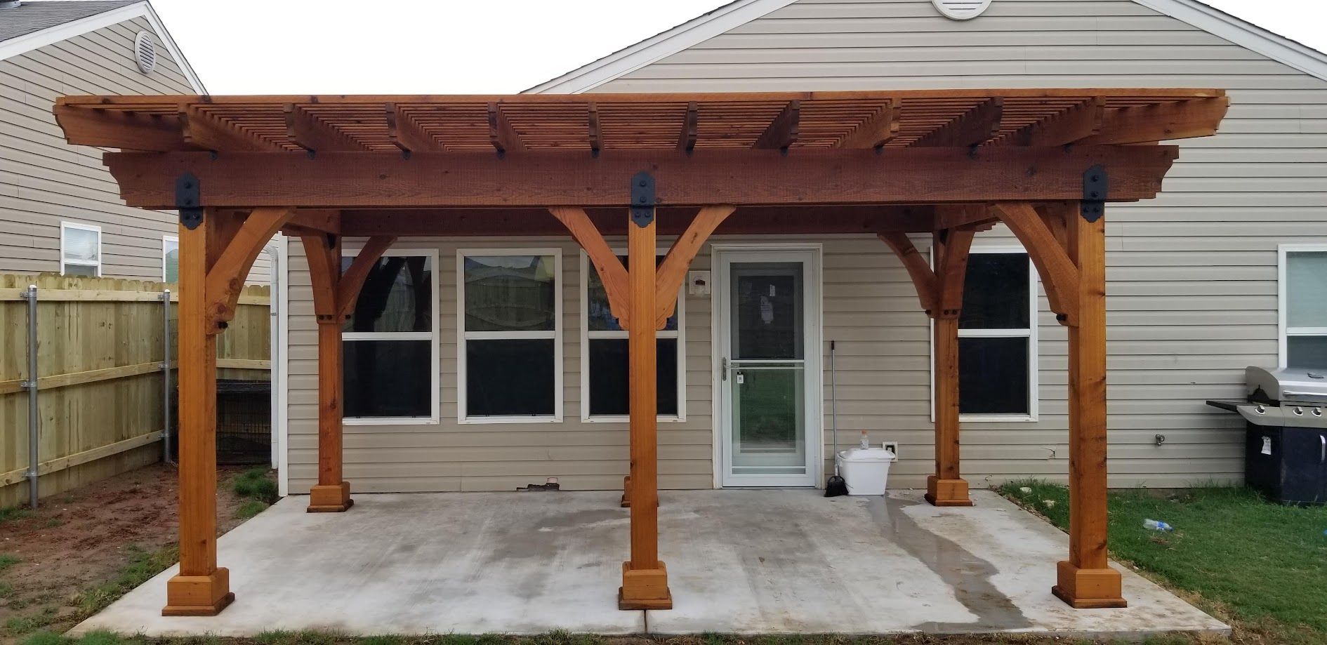 Wooden pergola covering a concrete patio behind a house, with four posts and a small potted plant.
