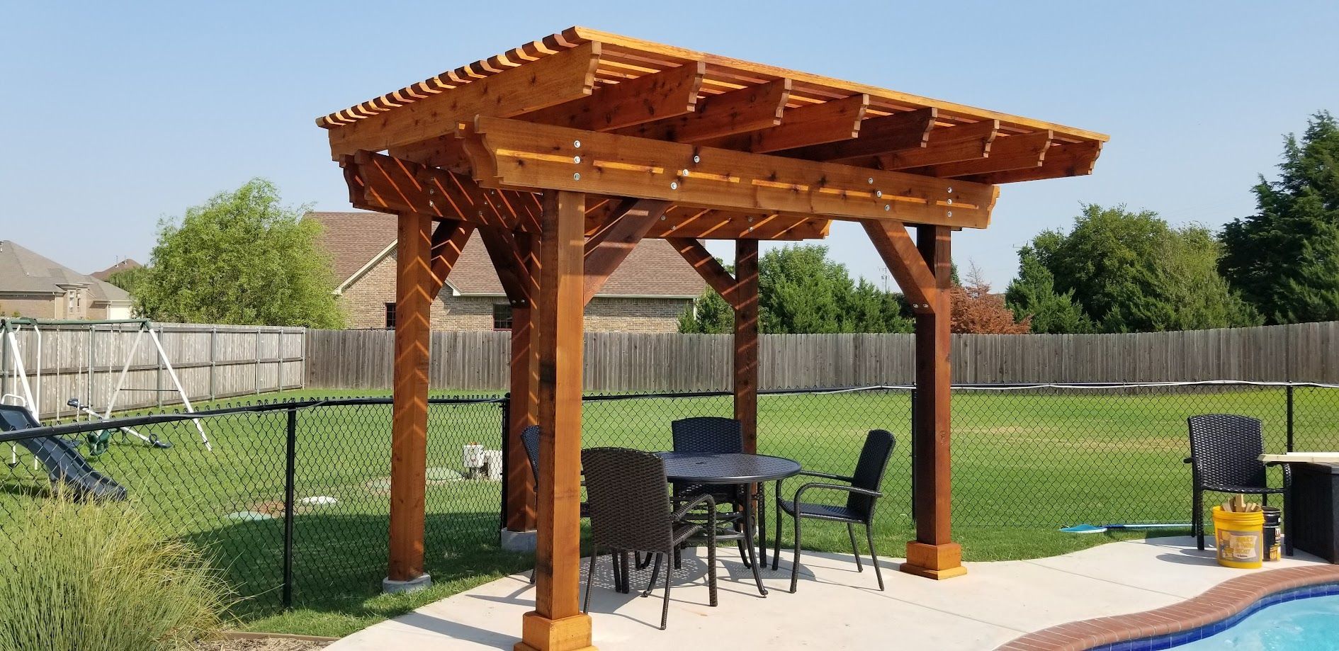 Wooden pergola shading a patio table and chairs in a fenced backyard, beside a pool.