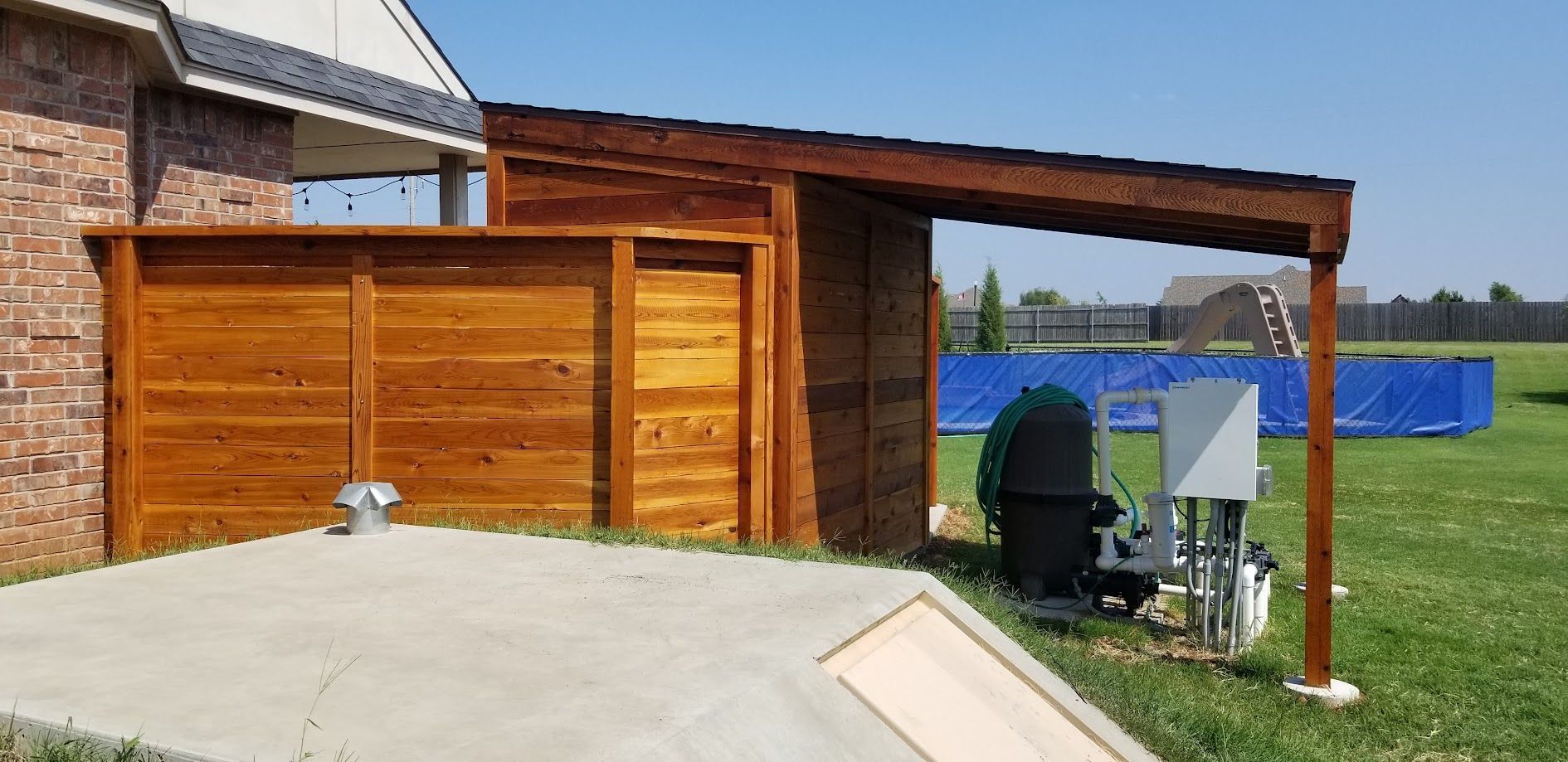 Wooden shed beside a sloped roof and outdoor utility equipment near a grassy field and blue barrier