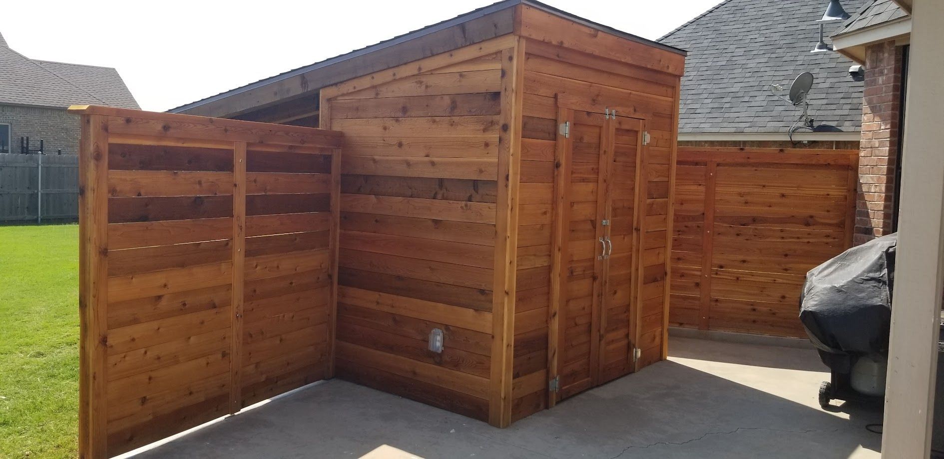 Wooden outdoor storage shed with slatted panels on a concrete patio near a stone wall