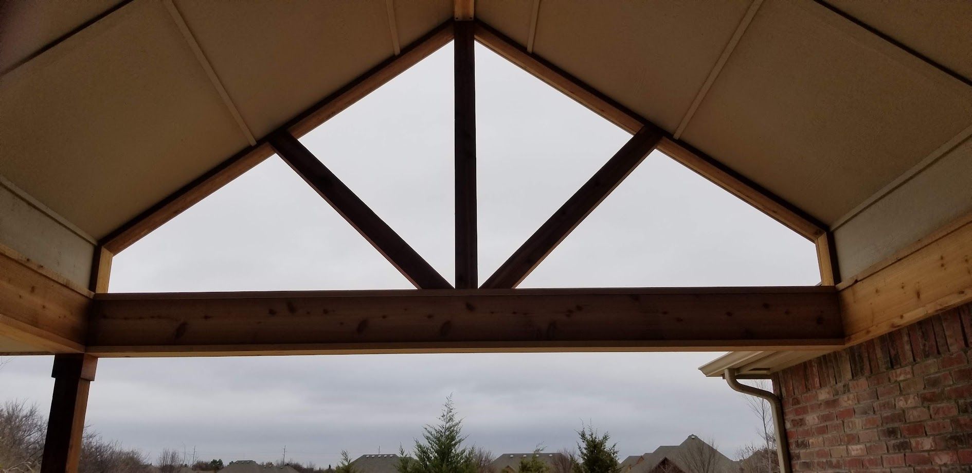 Open gazebo roof with wooden beams framing a cloudy sky and distant trees