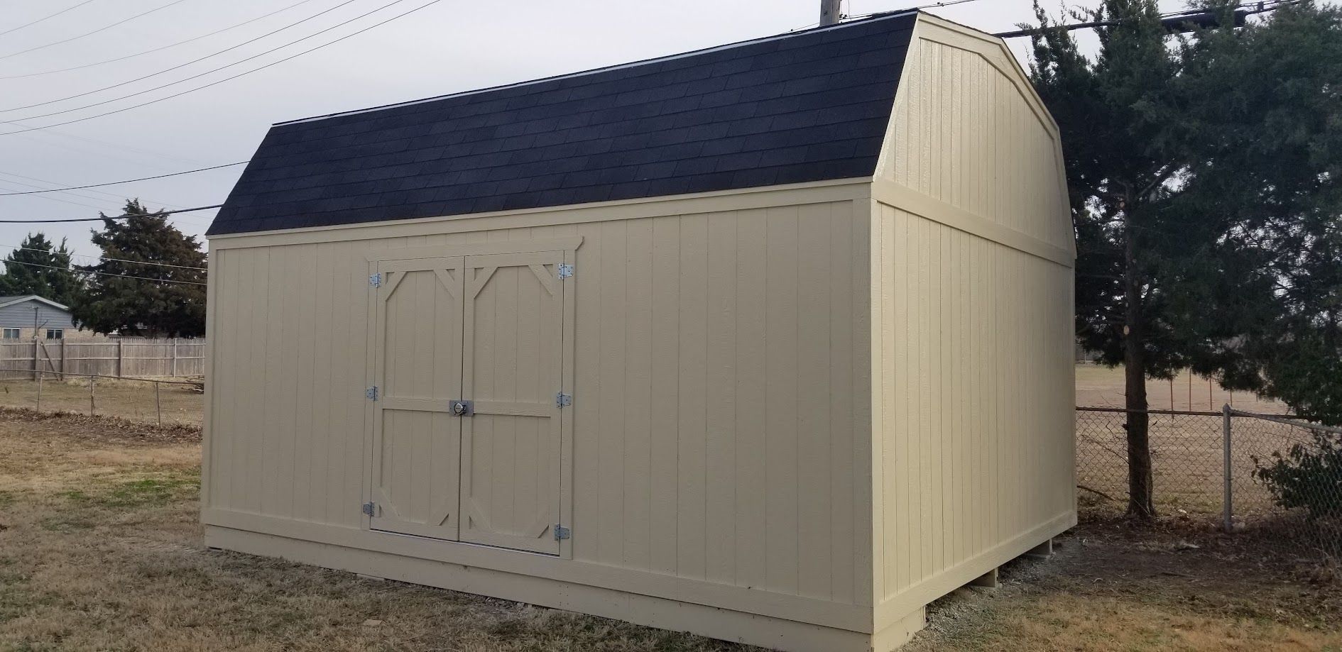 Beige metal shed with a dark roof sitting on a gravel yard near a fence and trees.